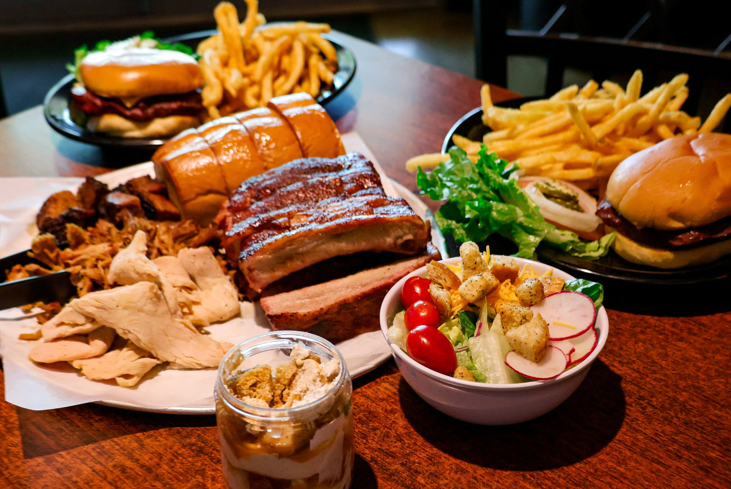 Diverse food items including burgers, fries, salad, ribs, smoked meat, and a dessert on a wooden table.
