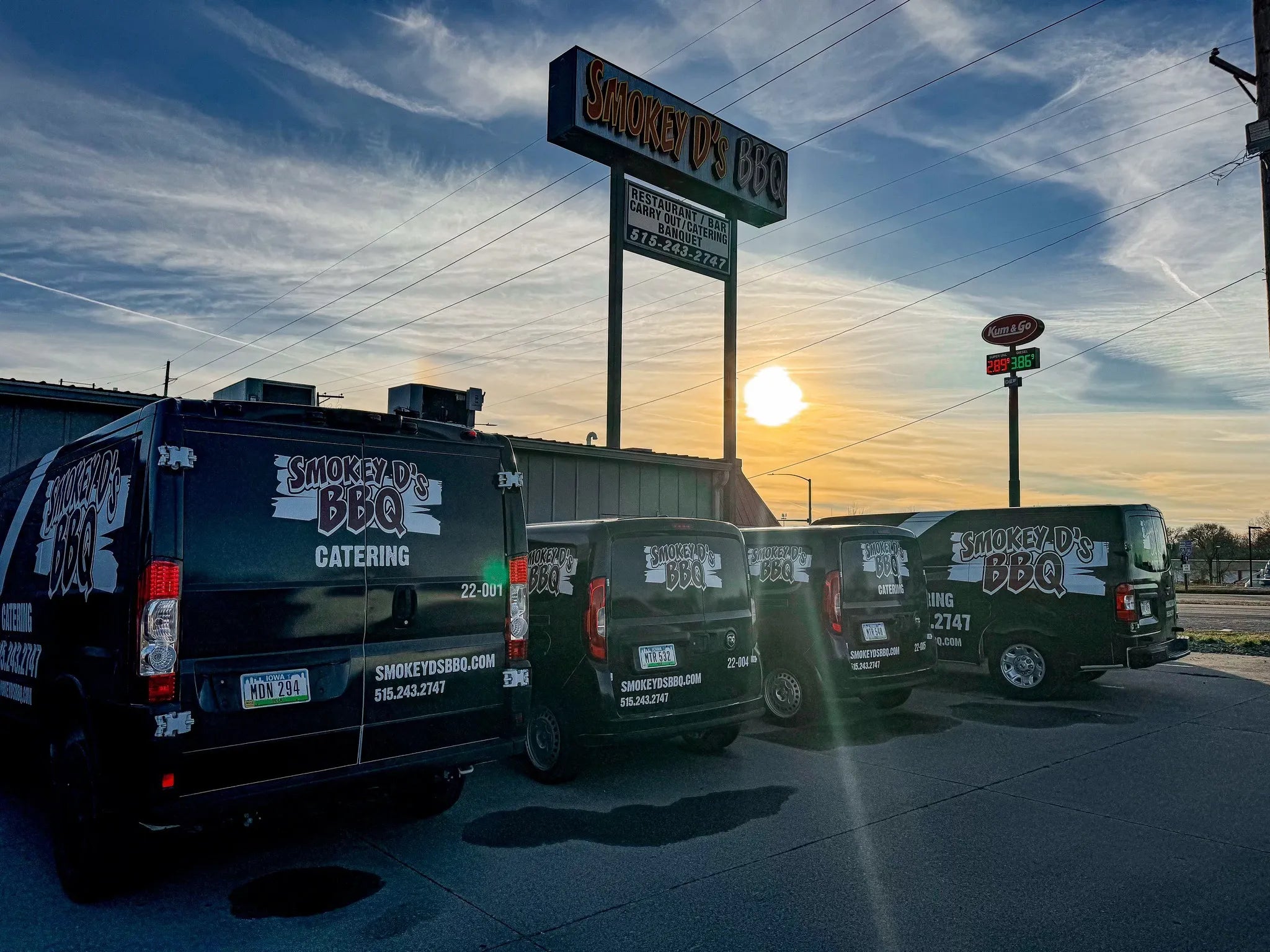 Three branded Smokey D's BBQ Catering vans parked in front of a restaurant with a sunset sky.