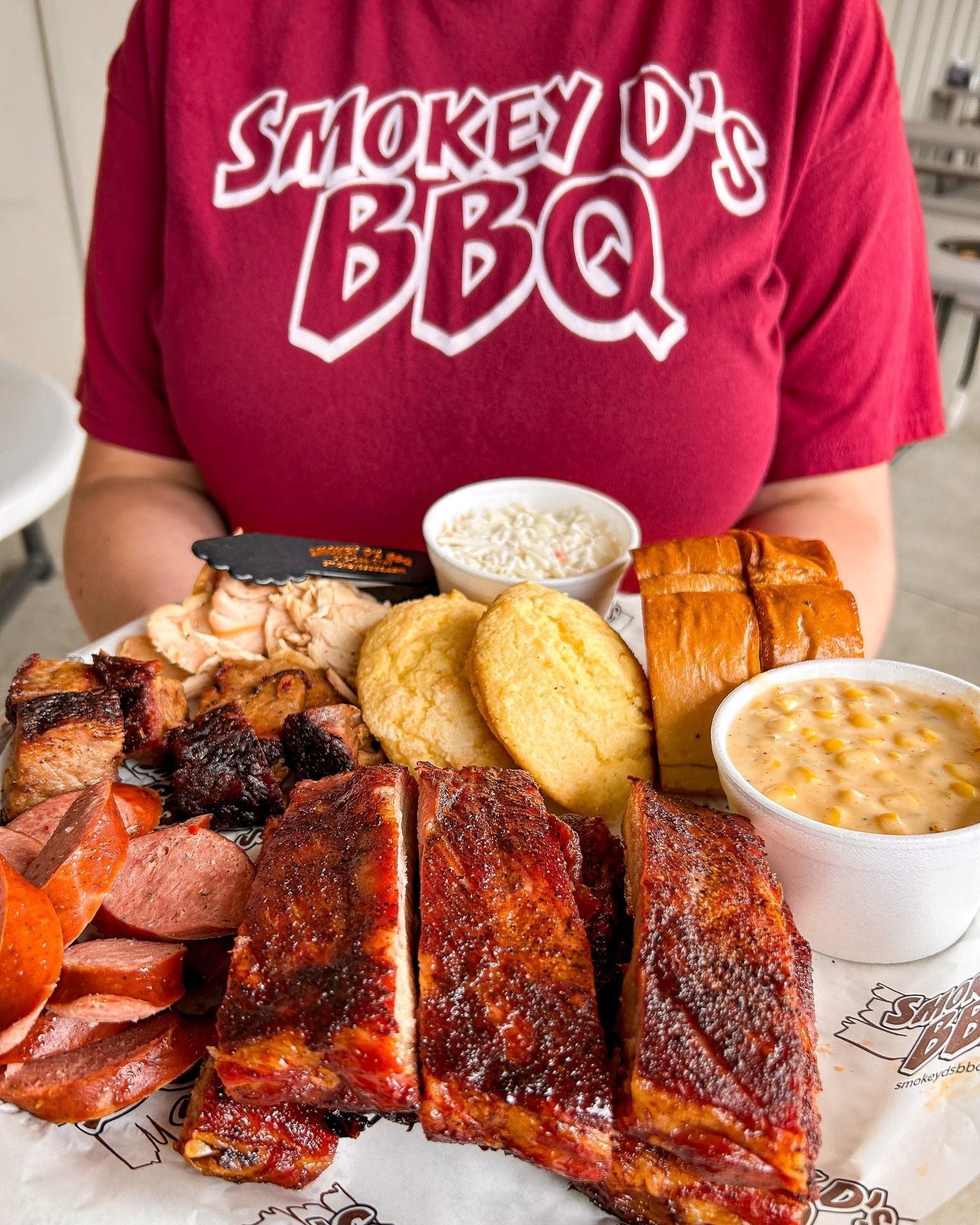 Person wearing a 'Smokey D's BBQ' shirt with a platter of barbecue meats, sides, and bread.