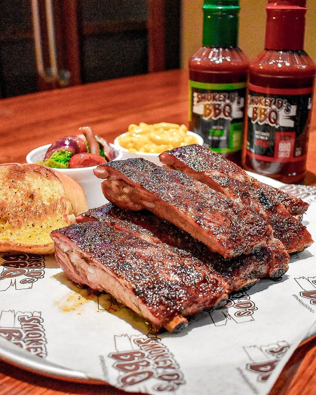 Platter of ribs with sides and bottles of barbecue sauce on a wooden table. 