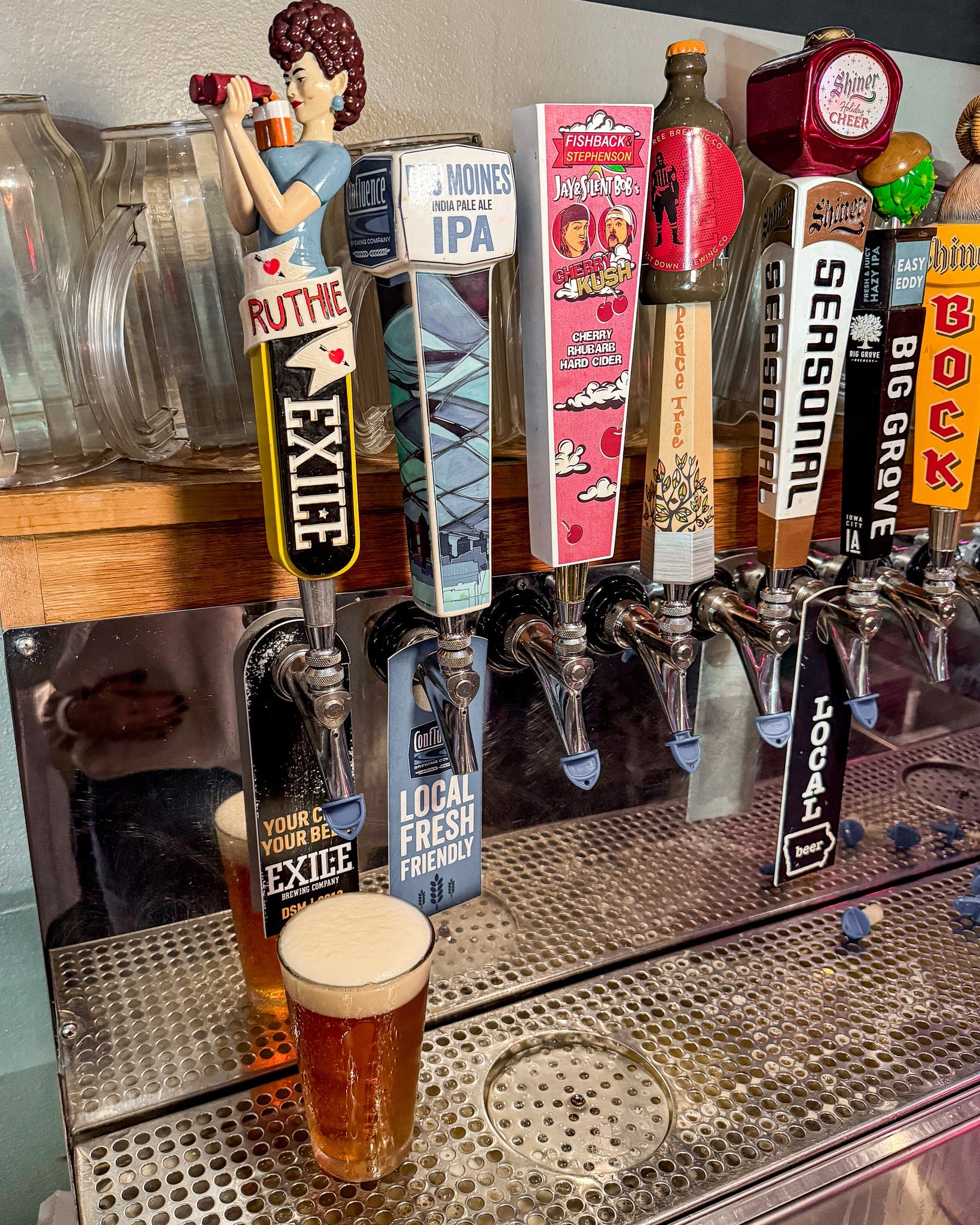 Beer taps with various labels at a bar, one tap pouring a glass of beer.
