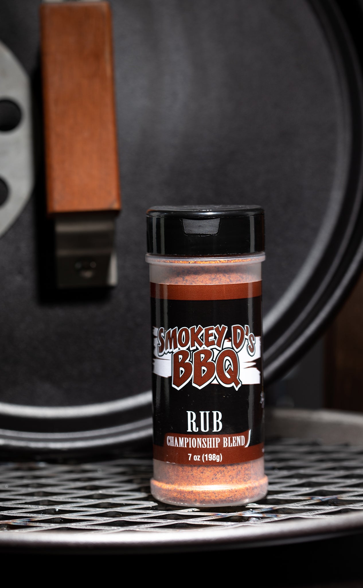 A 7 oz container of Championship Blend BBQ rub with a label that reads 'Smokey's BBQ RUB'. The container is placed on a grill.